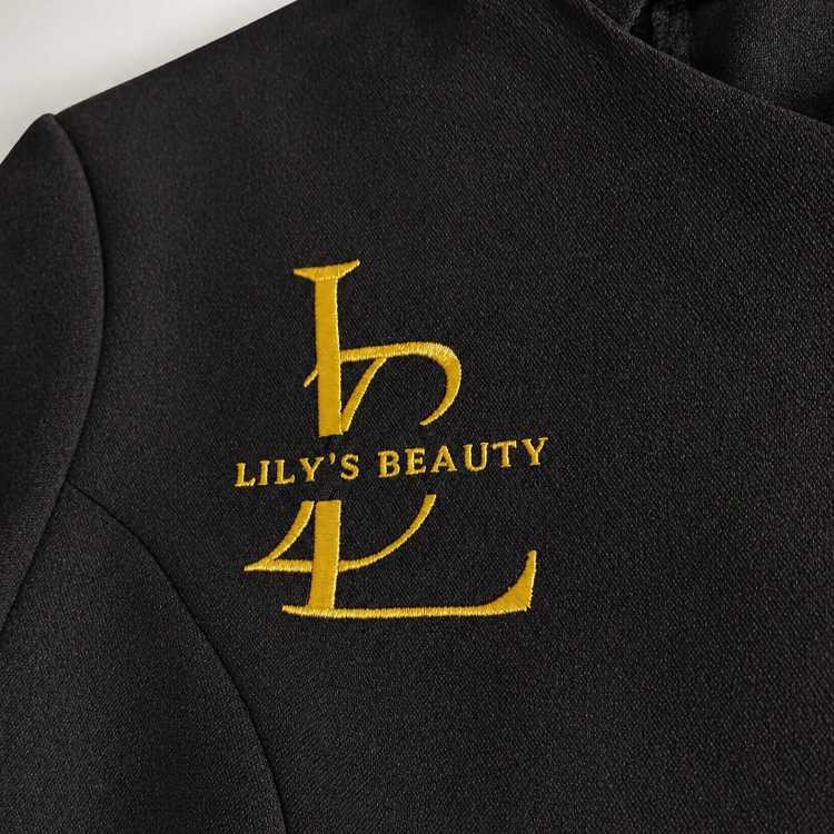 Beautician tunic – A customised black beauty salon tunic featuring elegant gold embroidered branding with the “Lily’s Beauty” logo on the chest, carefully finished with refined detailing, smooth fabric texture, and a professional luxury appearance.